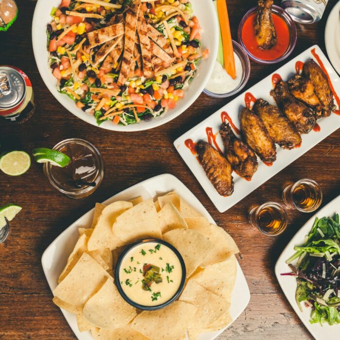 A table with colorful dishes, including a salad, chicken wings, a burger, nachos with dip, and drinks.