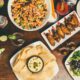 A table with colorful dishes, including a salad, chicken wings, a burger, nachos with dip, and drinks.