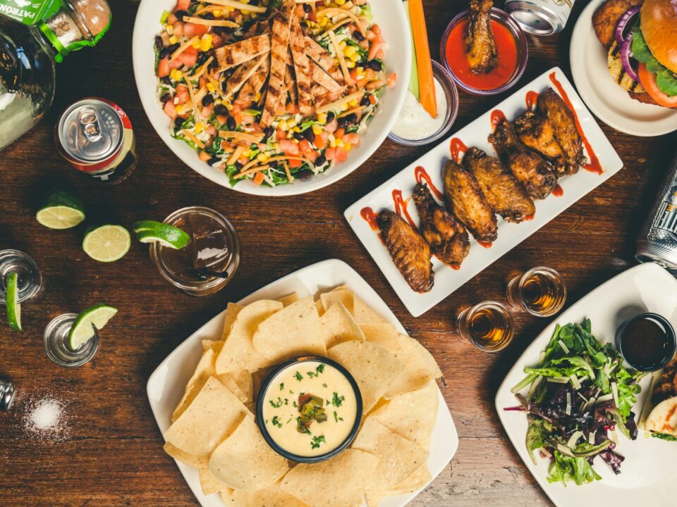 A table with colorful dishes, including a salad, chicken wings, a burger, nachos with dip, and drinks.