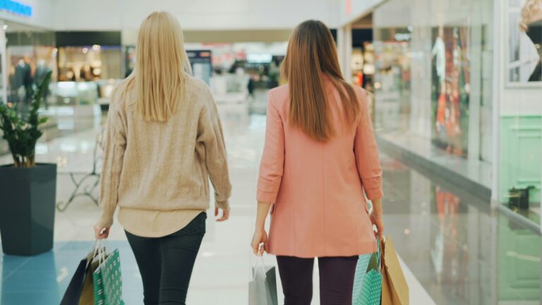 Rear shot of slender young women walking in shopping mall holding colorful paper bags and looking around at goods.